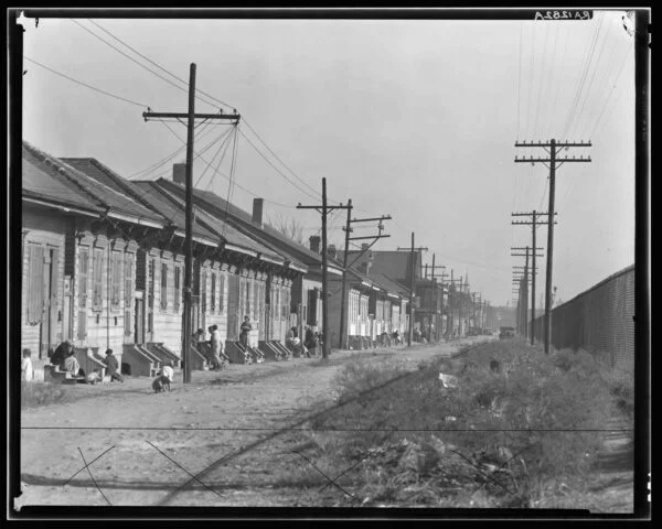 A black and white photograph of a row of single story houses with children and adults on the steps outside and overhead pylons above the dwellings. 