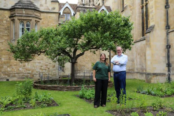A man and a woman standing on the grass among the flower beds and in front of an apple tree with buildings behind. 
