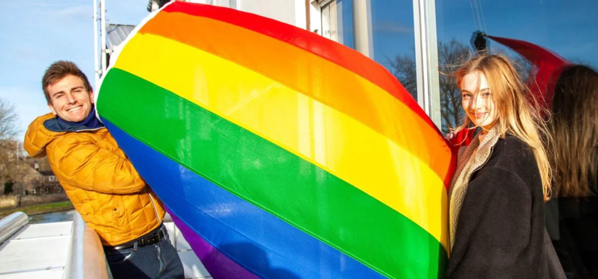 Two people holding the rainbow flag which is caught by the wind
