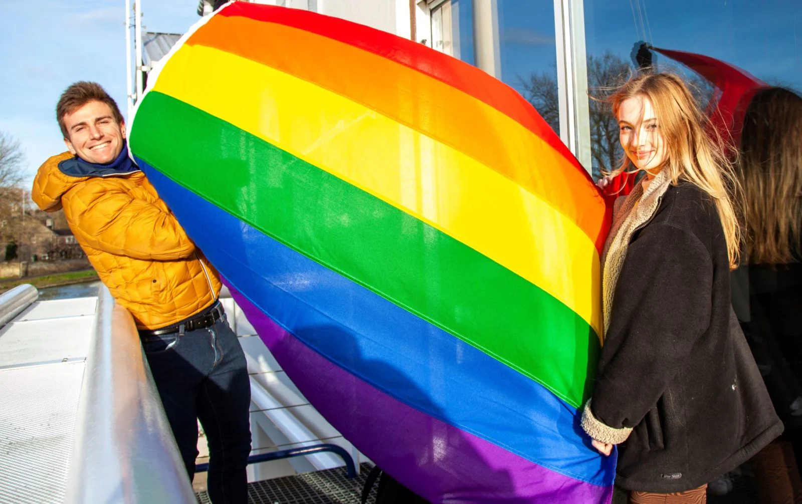Two people holding the rainbow flag which is caught by the wind