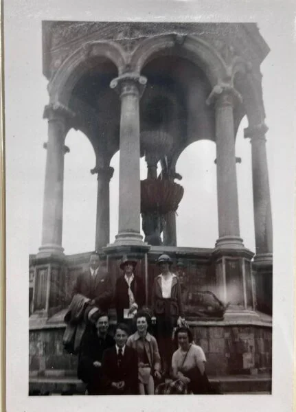 black and white image of men and women in two rows at the foot of an ornate stone fountain in Trinity College Cambridge. 